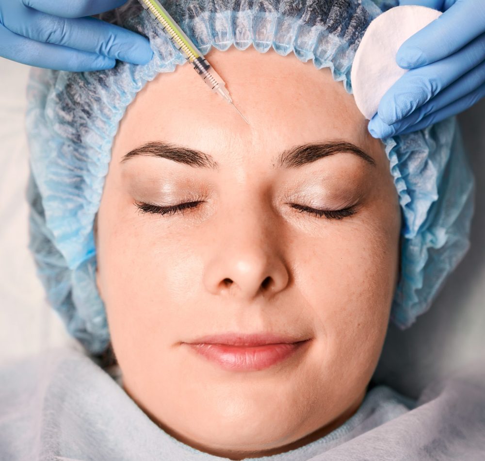 Close up of cosmetologist hands in sterile gloves making injection in young woman forehead. Female client in disposable shower hat receiving facial treatment. Concept of anti-aging procedure.